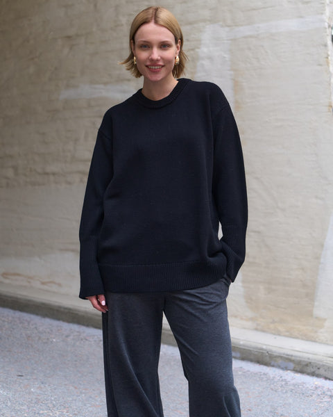 Person wearing a black sweater and gray pants standing against a beige wall.