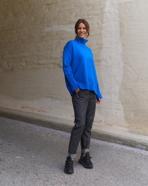 Person wearing a bright blue sweater and dark pants standing against a beige wall.