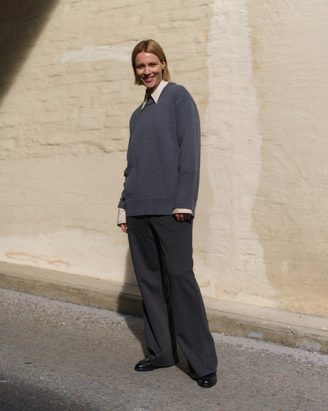 Person wearing a gray sweater and dark pants standing against a beige wall.