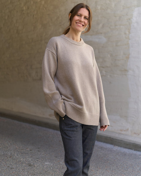 Woman wearing a beige sweater and dark pants standing against a plain wall.