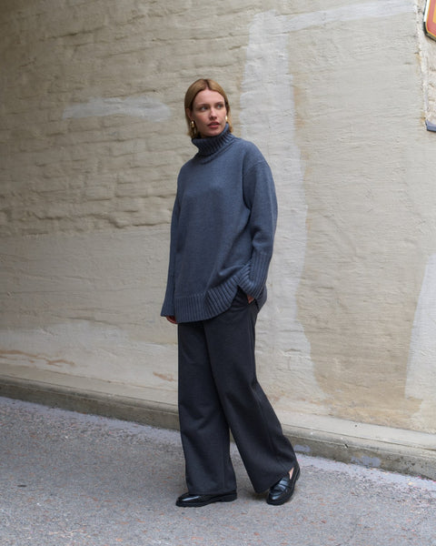 Woman wearing a gray sweater and black pants standing against a beige wall.