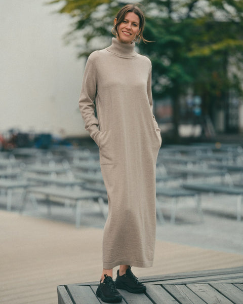 Woman wearing a long beige dress with a turtleneck standing outdoors.